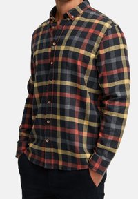 Homme portant une chemise en flanelle à manches longues boutonnée avec un motif à carreaux noir, rouge, jaune et gris, mains dans les poches.