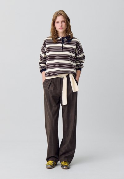 Polo a righe con maniche lunghe in crema e marrone, con colletto blu navy. Pantaloni marroni a righe allentati, legati con una cintura crema. Scarpe con dettagli gialli.