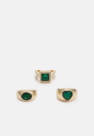 Três anéis de ouro com gemas verdes: um quadrado com realces claros, dois arredondados com formas ovais e de coração, todos com texturas lisas.