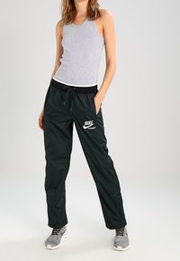 Person wearing a grey sleeveless top, black Nike drawstring pants, and grey sneakers, standing with hands in pockets against a plain background.