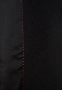 Gros plan d'un tissu noir avec une couture verticale de coutures rouges séparant un textile lisse et un matériau texturé.