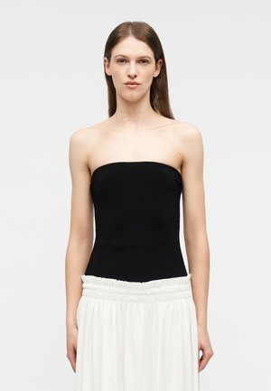 Kvinde med langt brunt hår iført en skulderfri sort top og en højtaljet hvid nederdel mod en ensartet lys baggrund.