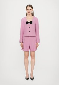 Pink jacket with black bow, four buttons, and long sleeves paired with matching shorts. Smooth texture and tailored fit. Black heels.