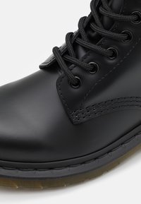 Musta nahkasaapas, jossa on sileä pinta, pyöreä kärki ja mustat kengännauhat. Varustettu metallisilla rei'illä ja tekstuurilla varustetulla kumipohjalla.