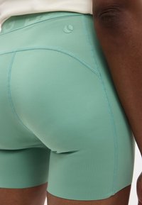 Άτομο που φοράει στενά αθλητικά σορτς μέντας με ορατές ραφές και ένα μικρό στρογγυλό λογότυπο κοντά στη μέση.