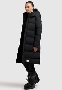Schwarze Daunenjacke mit einem langen, vertikalen Steppmuster, Kapuze und Reißverschluss. Hat ein Etikett am Saum und wird mit schwarzen Stiefeln kombiniert.