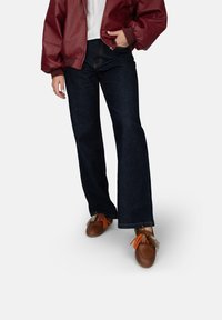 Bordeauxfarbene Bomberjacke, weißes T-Shirt, dunkelblaue Schlagjeans und braune Schuhe mit bunten Quasten. Glatte Texturen und lässige Passform.