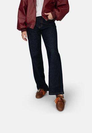 Chaqueta bomber color burdeos, camiseta blanca, jeans acampanados de mezclilla oscura y zapatos marrones con borlas de colores. Texturas suaves y ajuste casual.