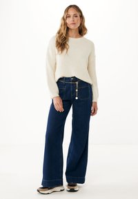 Sweter w kolorze kremowym, dziergany, z długimi rękawami, w połączeniu z ciemnoniebieskimi, szerokimi dżinsami z denimu, zdobionymi łańcuszkiem w talii.