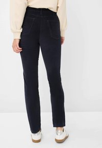 Donkerblauwe denim broek met een aangesloten ontwerp, hoge taille en twee achterzakken, gecombineerd met witte schoenen met metalen accenten.