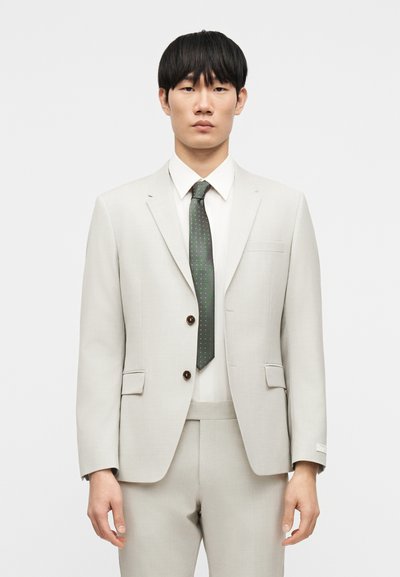 Ανοιχτό γκρι κοστούμι με δύο καφέ κουμπιά, λευκό πουκάμισο και πράσινη γραβάτα με μικρές λευκές βούλες. Ραμμένο κανονικό fit με πλαϊνές τσέπες. Ομαλή υφή υφάσματος.