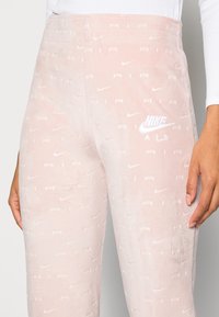 Rosa sammet joggers med ett texturerat mönster som visar upp upprepade "Nike"-logotyper och "AIR"-text, kompletterat med en broderad vit logodetalj.