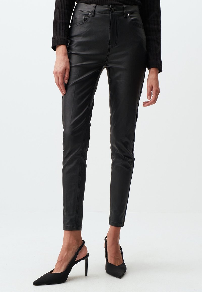 Pantalon en cuir noir slim fit porté avec un haut noir à manches longues et des escarpins slingback noirs à talons hauts sur fond blanc.