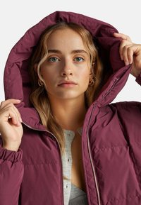 Burgundowa kurtka puchowa z kapturem, z metalowym zamkiem błyskawicznym i elastycznymi mankietami. Pod spodem widoczna szara koszula z guzikami. Gładka tekstura.