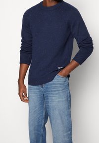 Man som bär en marinblåmelerad stickad tröja med ribbade muddar och nederkant, tillsammans med ljusblå jeans, med handen i fickan.