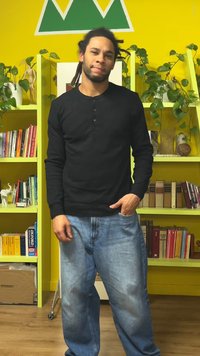 Svart henley-skjorte med lange ermer og knapper, kombinert med lyseblå, løstsittende denimjeans. Bakgrunnen har gule vegger og grønne hyller.