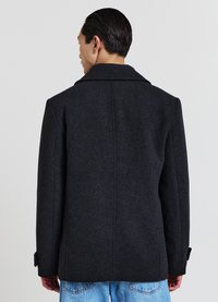 Manteau en laine gris foncé avec un col cranté, une coupe structurée et des détails de poignets à deux boutons, porté avec un pantalon en denim bleu clair.
