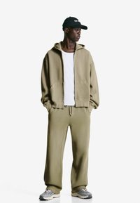 Khaki hoodie with front pockets and matching sweatpants, paired with a white T-shirt and gray sneakers, featuring a black cap with text.