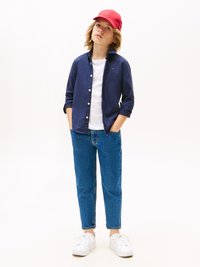 Marineblå knappskjorte over en hvit T-skjorte, kombinert med blå teksturerte jeans og hvite sneakers. En rød caps fullfører det avslappede antrekket.
