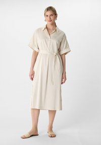 Beige Baumwollkleid mit einem Kragenhals, kurzen gefalteten Ärmeln, einer Taillenbindung und seitlichen Schlitzen. Getragen mit gewebten Rutsch-Sandalen.
