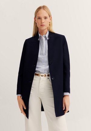 Navy coat with a minimalist design, featuring a round neckline, two buttons, and a tailored fit. Paired with a striped shirt and light pants.