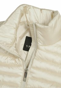 Lys beige dunjakke med quiltet tekstur og høj krave. Har en frontlynlås og en sort mærkeblæk indeni. Glat, skinnende materiale.