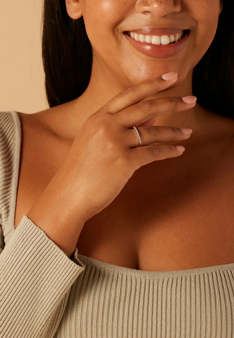 Silberner Ring mit kleinen klaren Steinen, gehalten von einer Hand mit mittlerem Hautton. Die Person trägt ein beige, geripptes Oberteil. Glatte Textur, dezenter Glanz.