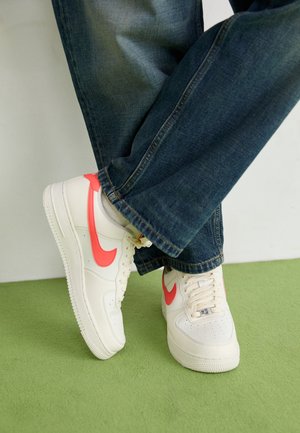 Persona che indossa sneaker bianche con loghi swoosh rossi e jeans blu scuro, in piedi su un tappeto verde chiaro con le gambe incrociate.