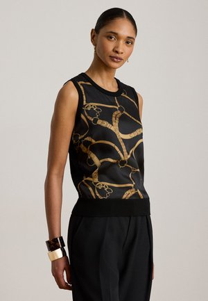 Vrouw draagt een zwarte mouwloze top met gouden kettingprint en zwarte hoge taillebroek, gecombineerd met stevige armbanden en gouden oorbellen.