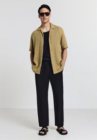 Uomo che indossa una maglietta a maniche corte in cotone oversize beige, una canotta nera, pantaloni neri con cordino, sandali neri e occhiali da sole rettangolari scuri, in piedi con le mani in tasca.