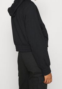 Black cropped hoodie with a relaxed fit, elastic cuffs, and a hood, made from soft cotton-like material. Features minimal detailing.