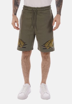 Uomo che indossa pantaloncini verde oliva con disegni di piume gialle sulle cosce, scarpe da ginnastica bianche e maglietta bianca, mostrando braccia tatuate.