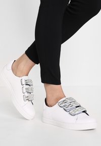Chaussures de sport blanches avec trois sangles métalliques à boucle, en cuir lisse, avec une semelle plate, assorties à un pantalon noir ajusté.