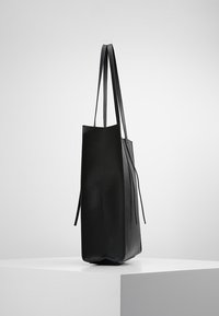 Grand sac fourre-tout en cuir noir avec des fines bretelles, se tenant debout sur un piédestal blanc contre un fond gris clair uni.