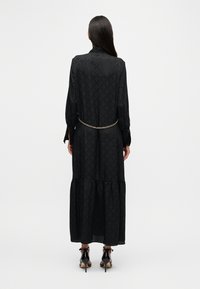 Robe maxi noire à manches longues, motif texturé, longueur sous le genou, taille cintrée avec accent en chaîne dorée, et ourlet évasé.