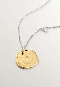 Colgante dorado con una "E" grabada, unido a una cadena de plata con un acento de cristal redondo. Acabado texturizado, forma circular.