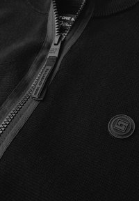 Svart zip-up tröja med strukturerad stickning, försedd med en slät dragkedja och en rund logotyp-patch på vänster sida.