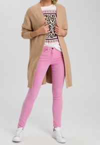 Cardigan lungo beige sopra una camicia bianca con design a stampa leopardo, abbinato a jeans skinny rosa pastello e sneakers bianche.