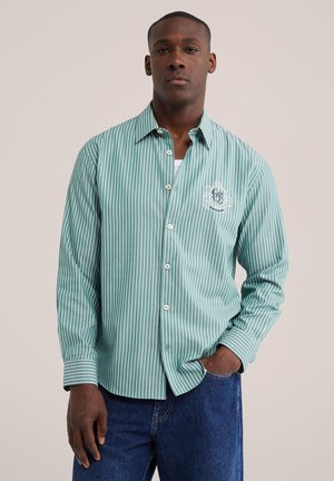 Mand iført grøn og hvid vertikalt striped knap-op skjorte med broderet logo, blå jeans, står med den ene hånd i lommen.