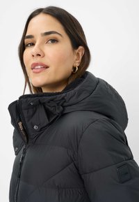 Jonge vrouw met donker haar, gekleed in een zwarte gewatteerde jas met hoge kraag en capuchon, die iets omhoog kijkt tegen een effen achtergrond.