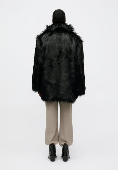 Osoba s krátkými vlasy v černém kabátu z umělé kožešiny, béžových kalhotách a černých botách, viděná zezadu na jednobarevném pozadí.