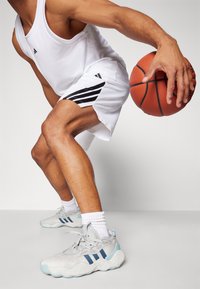 Homem a driblar uma bola de basquete, vestindo uma camisola sem mangas branca da Adidas, calções brancos com riscas pretas e sapatilhas Adidas cinza claro.