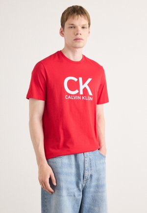 Crvena pamučna majica s okruglim izrezom s bijelim otisnutim logotipom "CK CALVIN KLEIN". Nosena uz svijetloplave traper šorts.