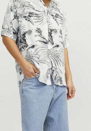 Personne portant une chemise blanche à manches courtes avec un imprimé feuilles tropicales noires et un jean bleu clair, une main dans la poche.