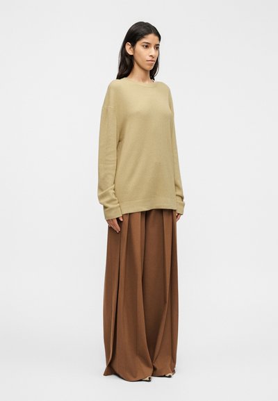 Pull beige en maille avec une coupe décontractée et des manches longues, porté avec un pantalon large marron qui tombe élégamment jusqu'au sol.