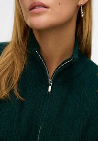 Temno zelena zip-up jopica z rebrasto teksturo in visokim ovratnikom; ima srebrno zadrgo in poudarjene šive.