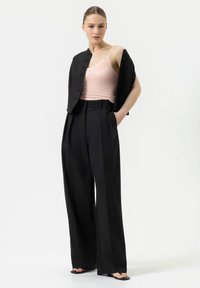 Czarne spodnie o wysokim stanie z zakładkami, zestawione z jasnoróżową koszulą na ramiączkach z koronkowymi akcentami. Modelka nosi czarną dopasowaną marynarkę.