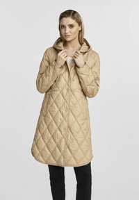 Beżowy płaszcz z kapturem o pikowanej fakturze i długiej sylwetce, z zatrzaskami oraz teksturowanym wzorem diamentowym. Idealny na chłodne dni.