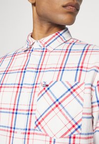 Närbild av en person som bär en vit rutig skjorta med röda och blå linjer och en knäppt krage, vilket visar en bröstficka.