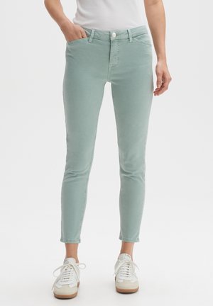 Osoba ubrana w jasne zielone dopasowane spodnie i białe trampki, stojąca z jedną ręką w kieszeni na jednolitym tle.